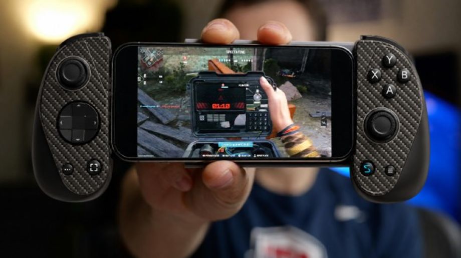 Close-up of a person holding a smartphone fitted with a black ergonomic mobile gaming controller while playing a first-person shooter game. The controller features textured grips, dual analog sticks, a D-pad, and ABXY buttons, with the gameplay screen in sharp focus against a softly blurred indoor background.