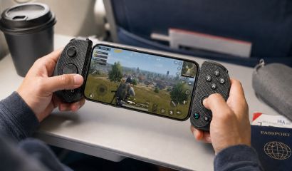 A person uses a smartphone attached to a USB-C mobile gaming controller while playing a battle royale game during a flight. The controller provides ergonomic console-style controls for comfortable gaming in an airplane seat, with a travel coffee cup and passport nearby, highlighting portable gaming on the go.